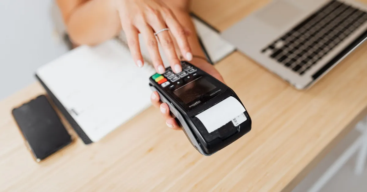 Close-up of a hand using a payment terminal beside a laptop on a wooden countertop.