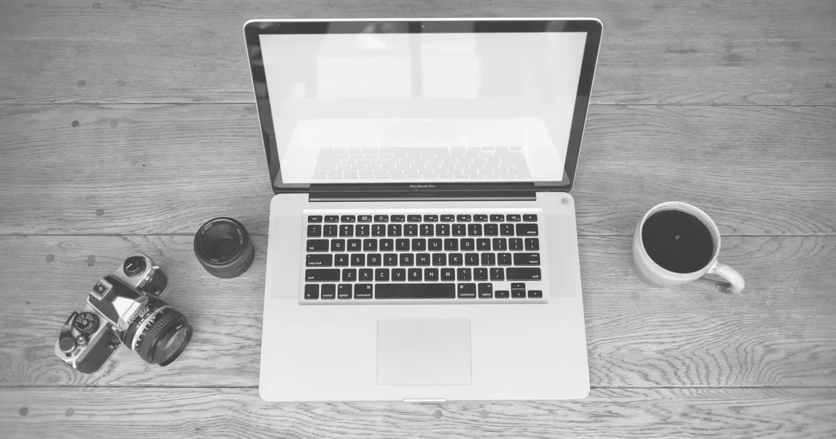Monochrome image of a laptop, camera, lens, and coffee cup on a wooden desk