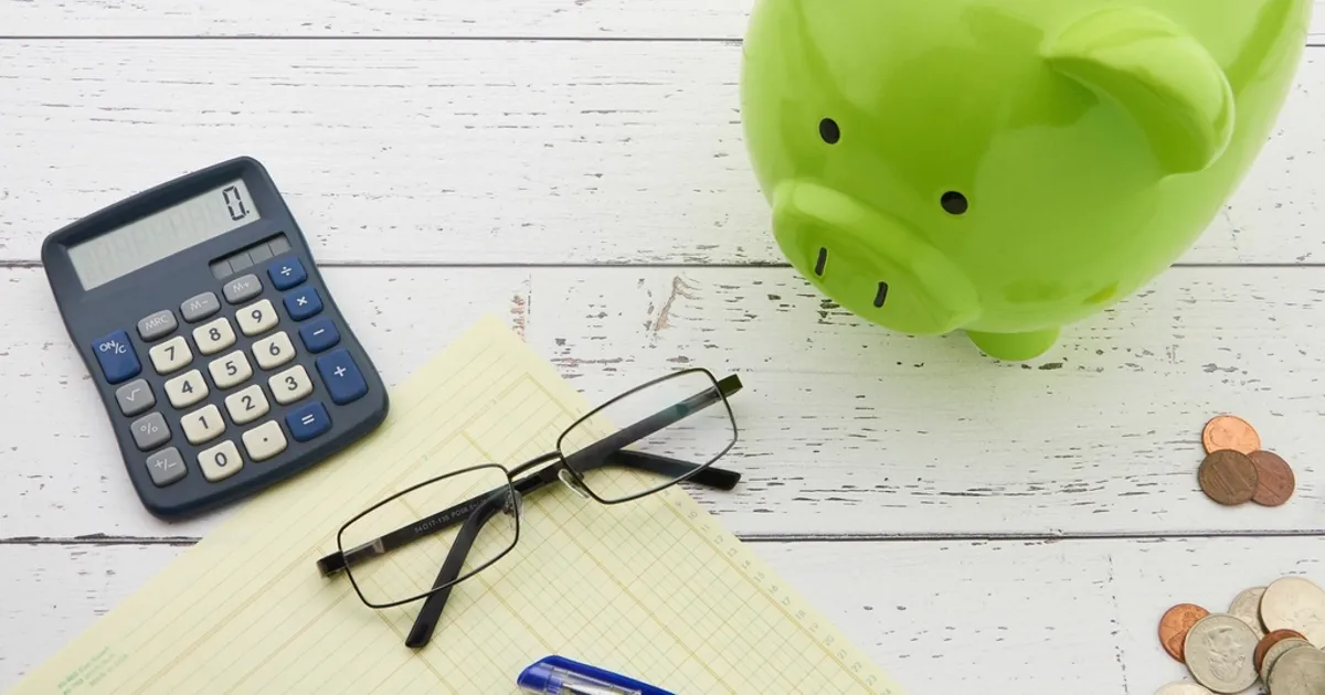 Cofrinho verde ao lado de uma calculadora, óculos e moedas sobre a mesa