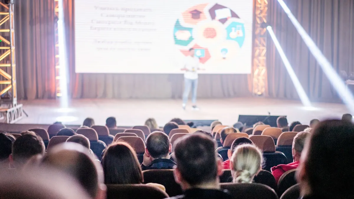 Audience attentively watching a presentation in a conference setting, with stage lights and speaker visible.