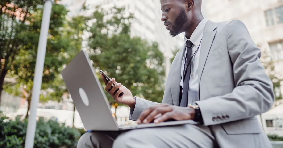 Businessman in a suit using a laptop and smartphone in an urban setting, blending technology with business.